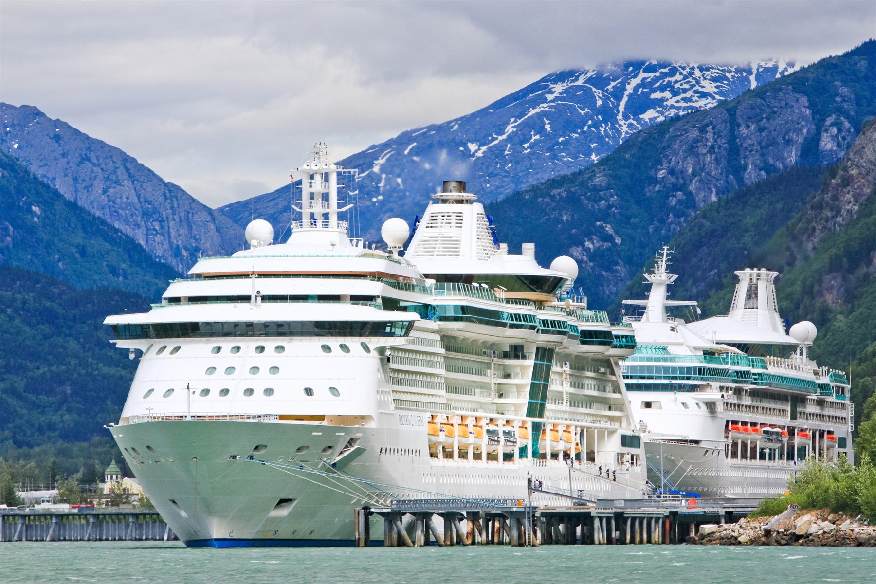 Two cruise ships docked side by side below forested, snow-streaked mountains